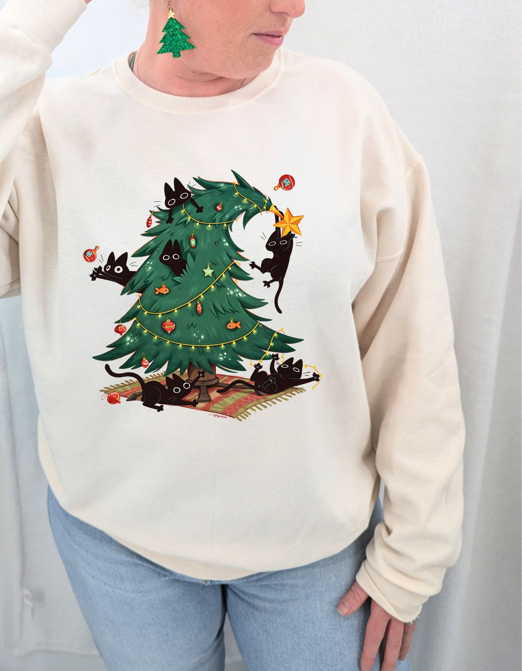 Person wearing a cream sweatshirt with a Christmas tree and cat design on a plain background