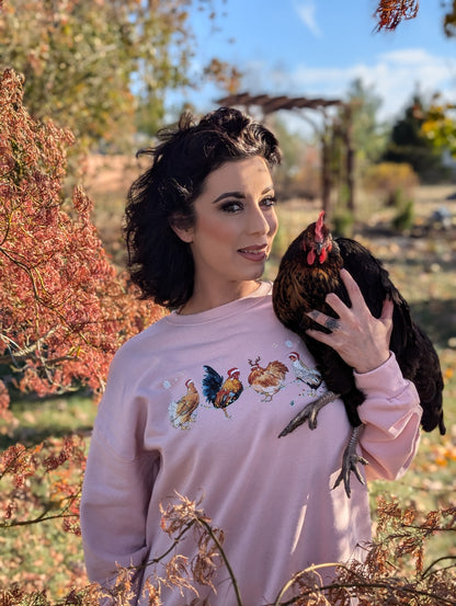 Woman wearing a pink sweatshirt with chicken designs, holding a black chicken outdoors.