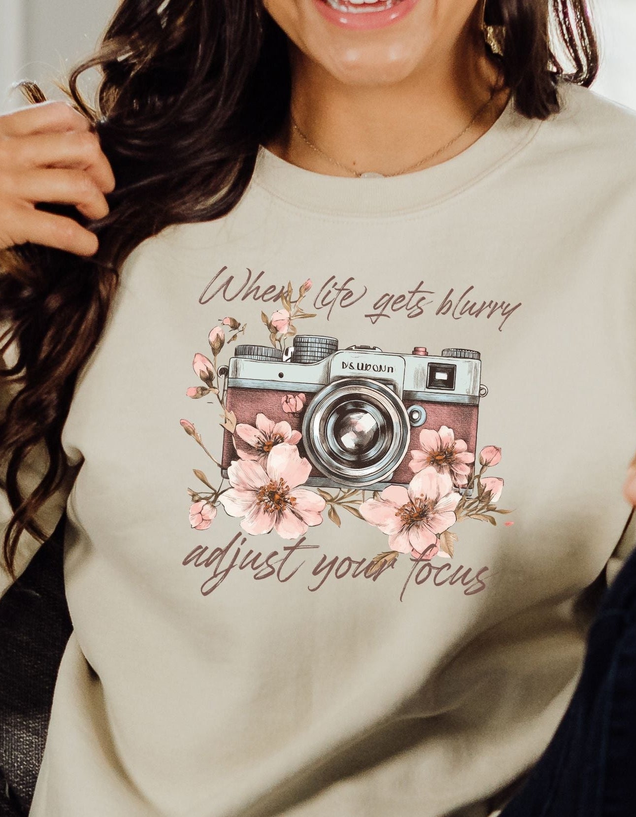 Person wearing a beige sweatshirt with a camera and floral design and text.